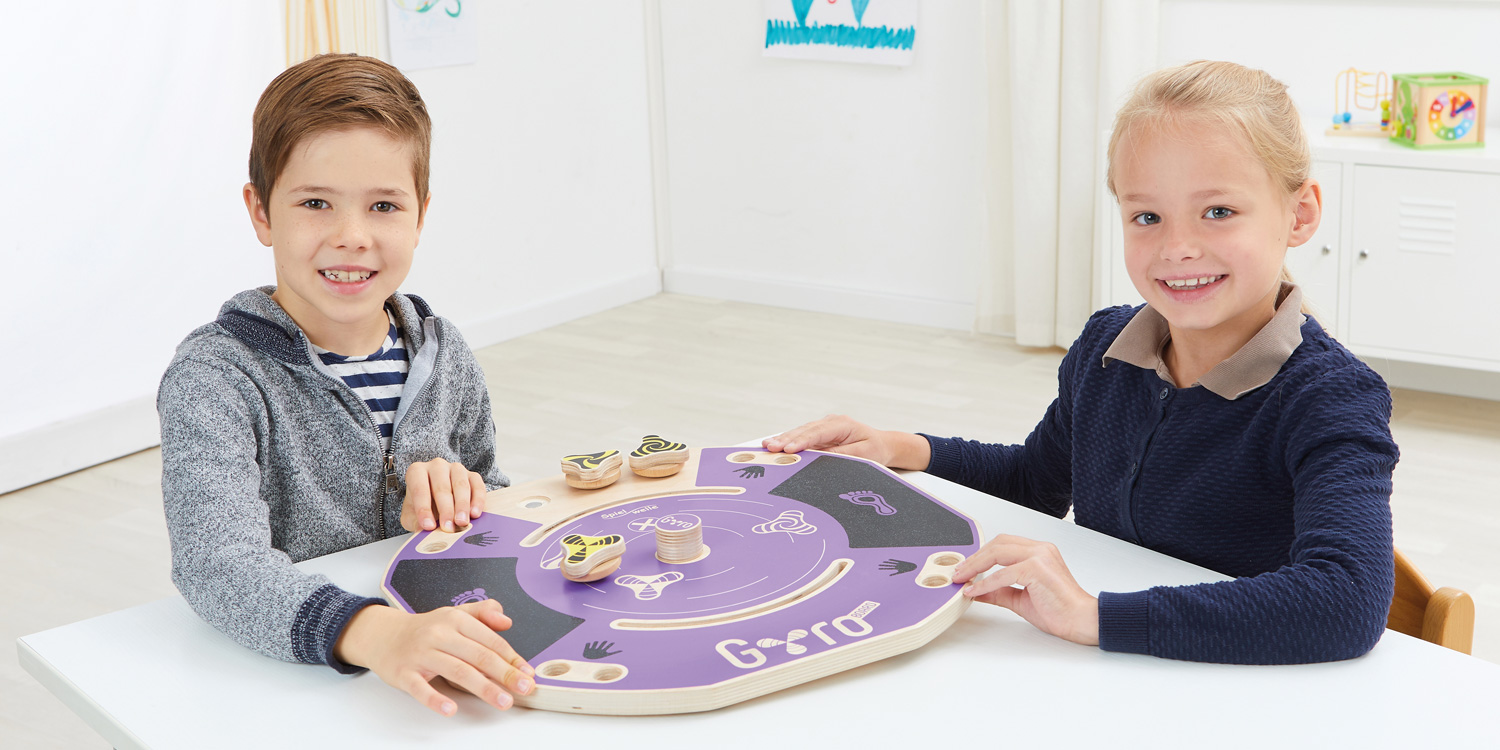 2 Kinder spielen mit dem Gyro-Board auf dem Tisch und bewegen die Spielscheibe mit den Grifflöchern.