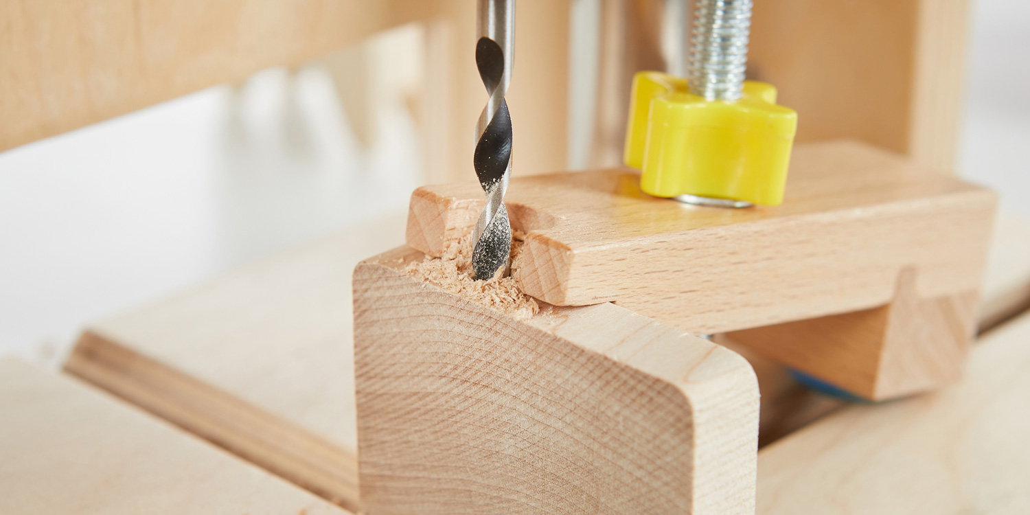 Ein Holzklötzchen ist fest auf dem Bohrtisch der Bohrmaschine eingespannt. Ein Holzbohrer dreht sich gerade und präzise in das Holz hinein.