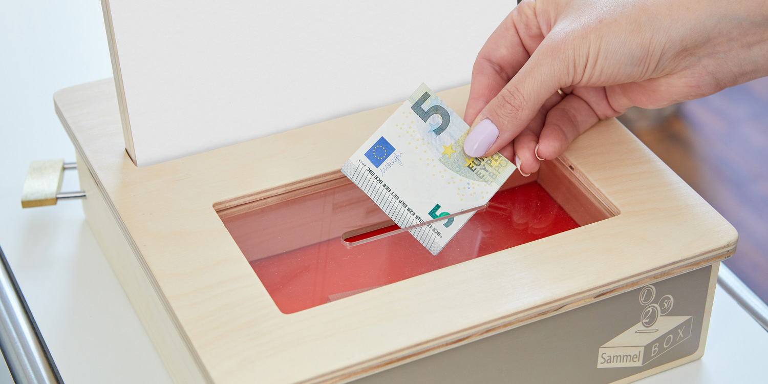 Eine Frau steckt einen Fünf-Euro-Schein in den Geldschlitz der SammelBOX.